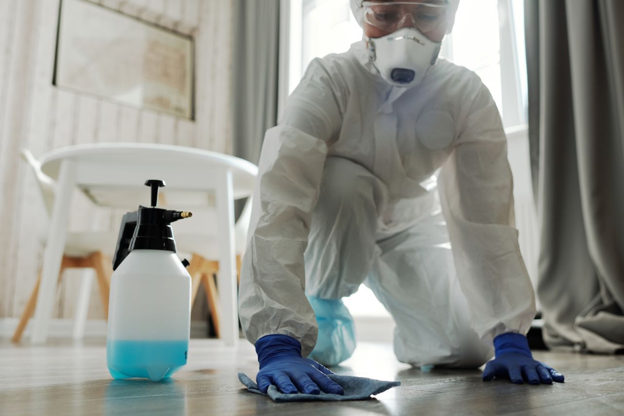 Une personne portant un équipement de protection nettoie un sol à l’intérieur à l’aide d’un spray désinfectant et d’un chiffon.